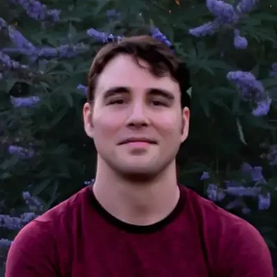Chiropractic Austin TX male massage therapist with brown hair wearing a maroon shirt standing in front of a lilac bush Meet The Team